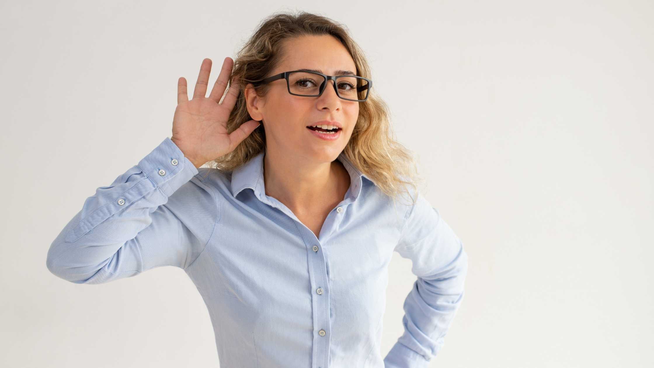 Young woman wearing glasses and a blue shirt holding hand to her ear against a grey background