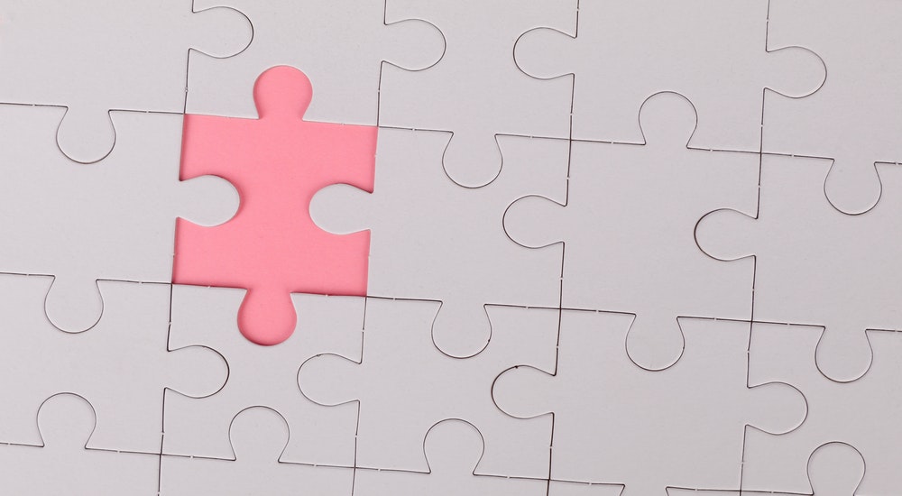 Close up of white jigsaw puzzle in assembled state with one piece missing revealing pink background